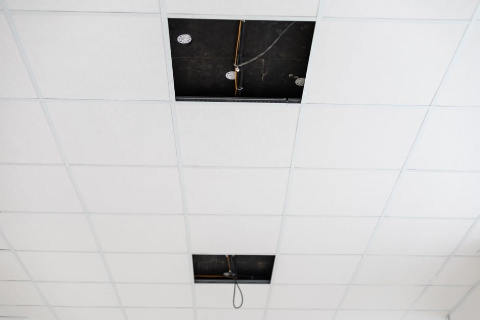 Suspended drop ceiling with two tiles removed, revealing the dark cavity, wiring, and pipes above the ceiling.