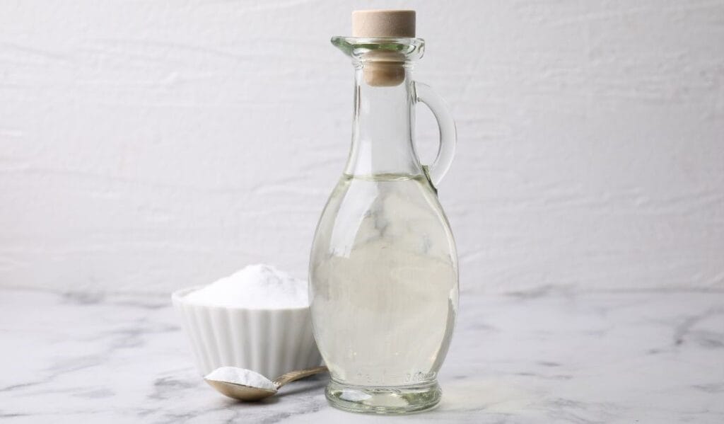 Bottle of vinegar next to a bowl and spoon of baking soda for drain cleaning.