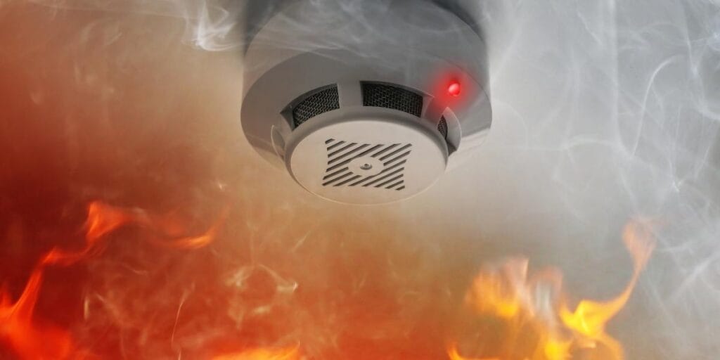 A ceiling-mounted smoke detector activated with a red light amidst thick smoke and fire.