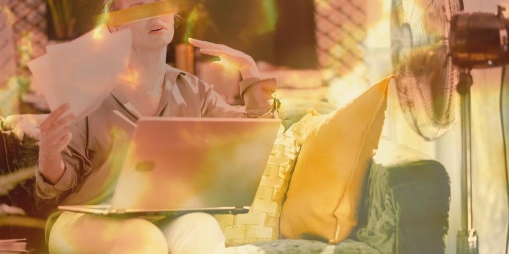 Woman fanning herself with paper while working on a laptop in a hot room with an electric fan.