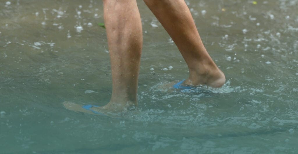 Close-up of a person's lower legs and feet wearing blue flip-flops, walking through murky, ripple-creating floodwater