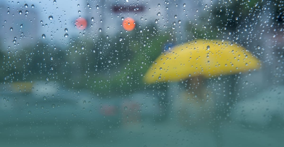 Raindrops on a window with a person holding a yellow umbrella for rainy season preparation